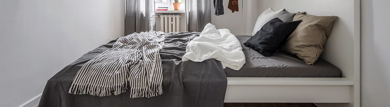 Imagem de quarto com paredes brancas e cama de solteiro com lençóis, travesseiros e cobertas cinzas.
