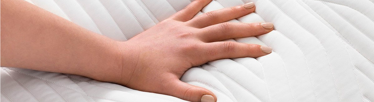 Imagem de mão caucasiana pressionando a superfície de um colchão branco para sentir a maciez.