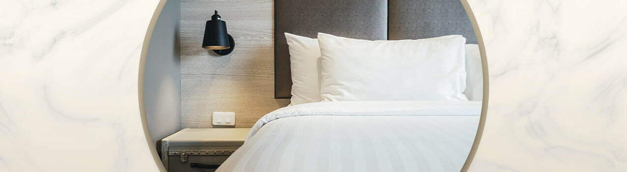 No centro da imagem, cama com cabeceira de casal cinza, travesseiros e cobertores brancos. No canto, uma luminária preta.